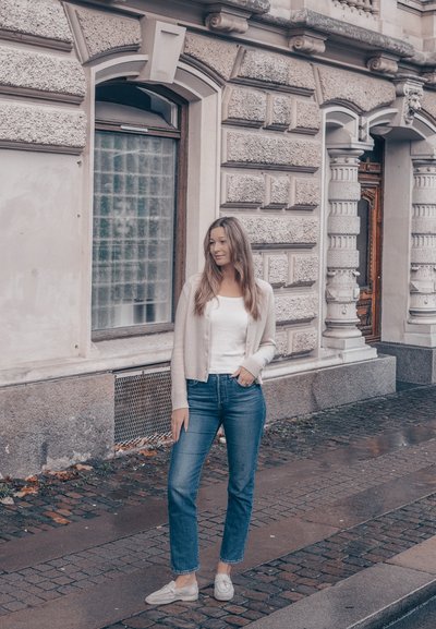 Cárdigan beige claro sobre una camiseta blanca, combinado con jeans azules y zapatos tipo slip-on gris claro. De pie sobre un camino adoquinado texturizado junto a un edificio histórico.