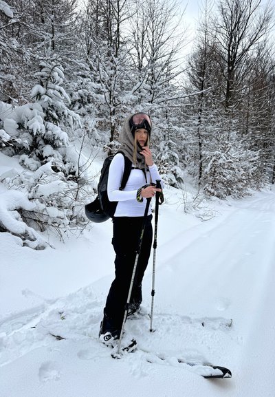 Persona de pie sobre esquís cubiertos de nieve en un bosque nevado, vistiendo ropa de invierno, sosteniendo bastones de esquí y llevando una mochila con un casco adjunto.