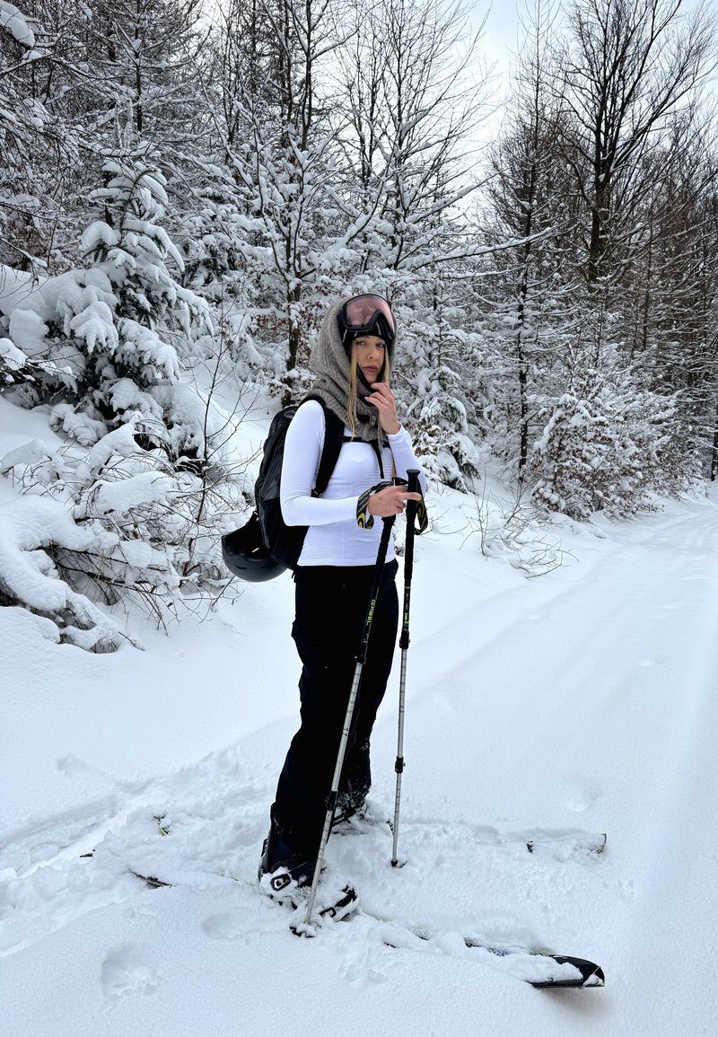 Personne debout sur des skis recouverts de neige dans une forêt enneigée, portant des vêtements d'hiver, tenant des bâtons de ski et portant un sac à dos avec un casque attaché.