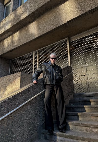 Chaqueta de cuero negra con detalles de flecos, acentos de botones y pantalones anchos negros. Escaleras de concreto y una barrera metálica enrejada en el fondo.
