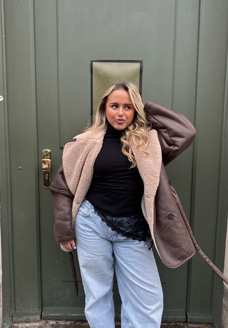 Jonge vrouw met lang blond haar die poseert bij een groene deur, gekleed in een bruine schapenvachtjas, een zwarte coltrui en lichtblauwe spijkerbroek.