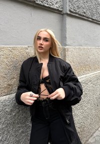 Czarna kurtka bombowa z ściągaczami, noszona na koronkowej bluzce. Model trzyma okulary, stojąc przed teksturowaną kamienną ścianą.