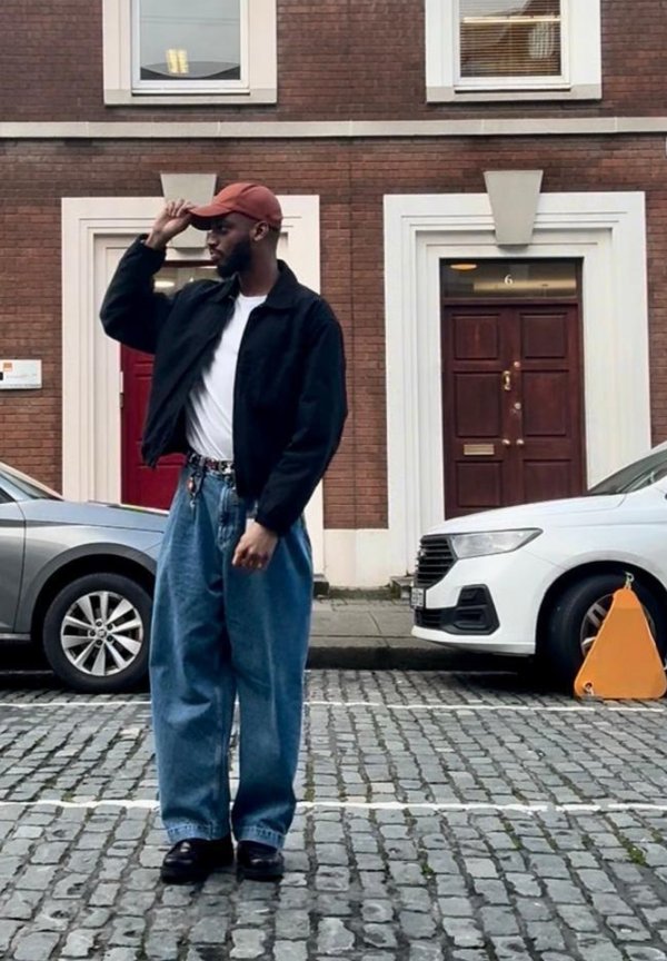 Man wearing a red cap, black jacket, white shirt, and loose-fitting jeans stands on a cobblestone street between parked cars in front of brick buildings.