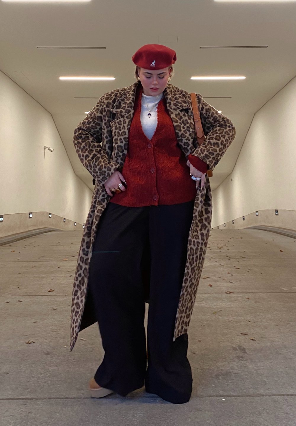 Καμηλοπάρδαλη καμπαρντίνα, κόκκινο πλεκτό γιλέκο, λευκό πουκάμισο, μαύρο παντελόνι σε φαρδιά γραμμή, κόκκινο μπερέ. Αξεσουάρ περιλαμβάνουν δαχτυλίδια και καφέ τσάντα ώμου.