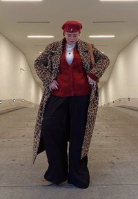 Καμηλοπάρδαλη καμπαρντίνα, κόκκινο πλεκτό γιλέκο, λευκό πουκάμισο, μαύρο παντελόνι σε φαρδιά γραμμή, κόκκινο μπερέ. Αξεσουάρ περιλαμβάνουν δαχτυλίδια και καφέ τσάντα ώμου.