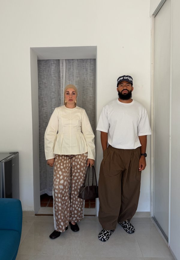 Un hombre y una mujer están de pie lado a lado en el interior, ambos llevan pantalones marrones con estampado sueltos y camisetas blancas, con expresiones faciales neutrales.