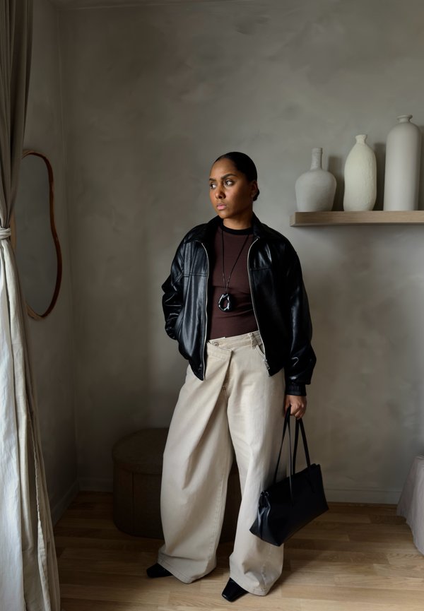 Ženska z črno usnjeno jakno, rjavo majico in širokimi bež hlačami, ki drži črno torbico in stoji v minimalistični sobi z vazami.