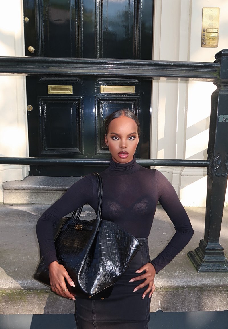 Femme en col roulé noir transparent et jupe, debout contre une rampe de porte noire, tenant un grand sac à main noir texturé sur un trottoir en ville.