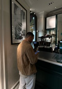Scène de café avec un homme portant une veste en daim beige utilisant un smartphone. Équipements de café verts et une œuvre d'art en noir et blanc visible.