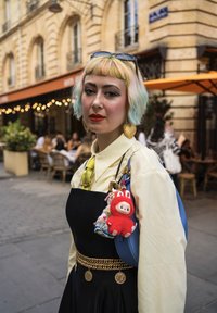 Joven con pelo corto multicolor, maquillaje audaz, pendientes en forma de corazón dorados, vestido negro sobre una camisa amarilla y una bolsa azul con un juguete de peluche rojo al aire libre.