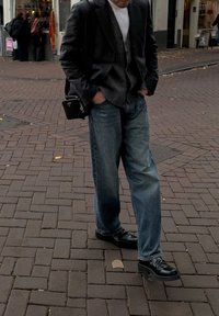 Sapatos de couro preto, jeans de ganga azul, camisola cinzenta e blazer preto. Uma pequena mala preta de ombro pendura-se da cintura. Calçada de pedra.