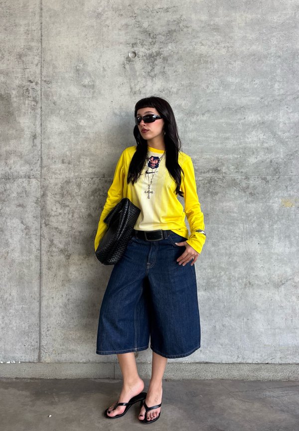 Mujer con camiseta amarilla de manga larga, pantalones cortos vaqueros anchos de pierna oscura, sandalias negras, gafas de sol y llevando un bolso de mano negro texturizado frente a una pared de hormigón.