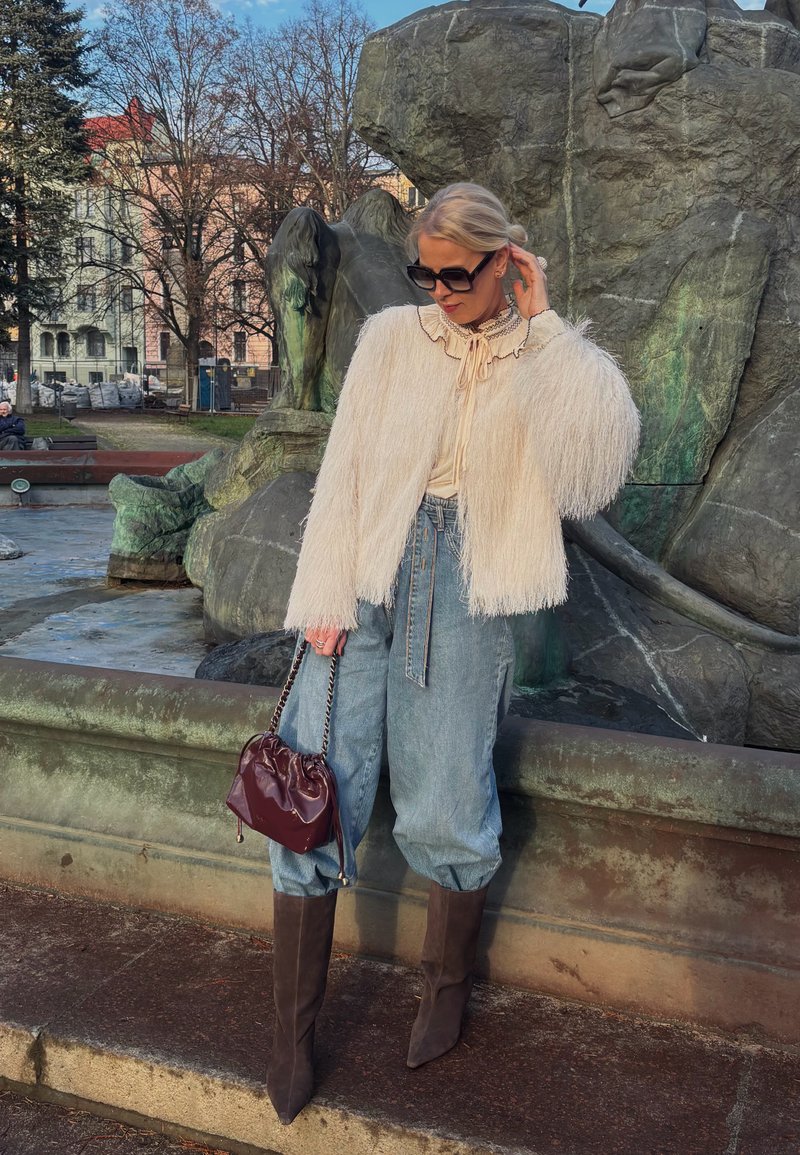 Veste ivoire à franges, blouse crème avec col à volants, jean bleu clair taille haute, bottes marron montantes jusqu'aux genoux, et sac à main bordeaux.