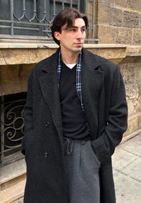 Jeune homme portant un manteau noir, un pull noir avec un sous-pull blanc et un pantalon gris, debout près d'un mur en pierre avec des détails en fer forgé.