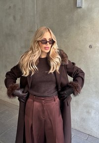 Manteau en laine marron avec poignets bordés de fourrure, pull marron foncé et pantalon bordeaux taille haute. Accessoires comprennent des lunettes de soleil élégantes et des gants.