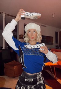 Biele vysoké podpätky s čiernymi polmesiacovými vzormi držané nad osobou v modrom svetri, čiernej vzorovanej sukni a bielej barete.