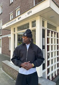 Schwarze Bomberjacke mit Reißverschluss, gestreiftes Hemd darunter, schwarze Hose, Accessoires bestehen aus einer Mütze und einer Brille, steht draußen vor einem Gebäude.