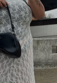 Balta nėrinių suknelė su raštuota tekstūra, juodas kreivai formos rankinė su petnešėle, laikoma ranka. Fone matomi betono elementai.