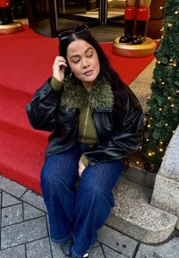 Jachetă neagră din piele cu guler din blană artificială verde, pulover verde închis, blugi largi albaștri și pantofi negri pe un fundal de covor roșu.