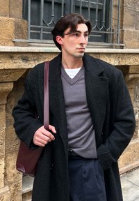 Νεαρός άνδρας που φοράει μαύρο παλτό και γκρι πουλόβερ με μπορντό τσάντα στον ώμο, στέκεται δίπλα σε πέτρινο τοίχο με παράθυρο με κάγκελα.