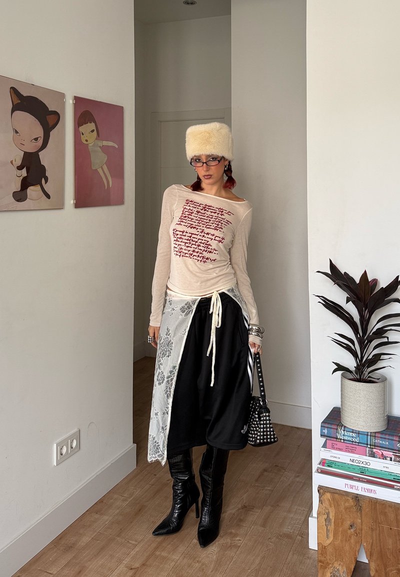 Haut crème à manches longues avec texte rouge, jupe noire, superposition en dentelle blanche, bottes en cuir noir montantes jusqu'aux genoux et chapeau blanc duveteux.