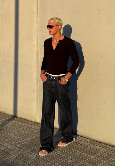 Camisa negra de manga larga con botones, jeans de mezclilla oscura y chanclas claras. Lleva unas gafas de sol grandes y está de pie contra una pared de concreto.