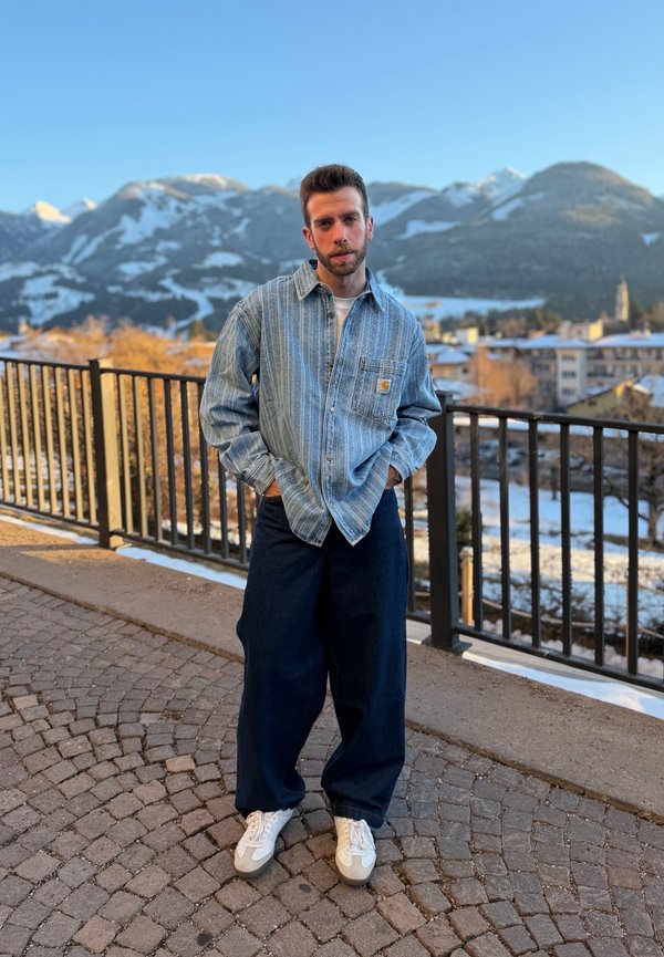 Hombre con barba vestido con camisa azul a rayas y pantalones anchos oscuros, de pie sobre un camino de piedra con montañas cubiertas de nieve al fondo.
