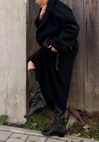 Zwarte wollen jas met oversized revers, gecombineerd met een zwarte asymmetrische rok en kniehoge zwarte leren laarzen. Geweven achtergrond.