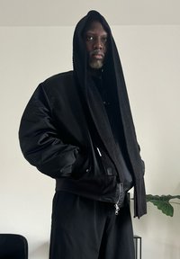 Schwarze Bomberjacke mit geripptem schwarzem Kapuzenpullover, langen Ärmeln und Reißverschluss. Über einem dunklen Hemd und schwarzen Hosen getragen. Einfaches, elegantes Design.