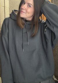 Temno siva prevelika jopa s kapuco z vrvico za zategovanje in srebrno kovinsko opremo. Mehka tekstura, veliki žepi na sprednji strani in izvezen logotip na prsih.