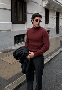 Άνδρας που φοράει γυαλιά ηλίου και μπορντό ριμπ ζιβάγκο κρατάει ένα μαύρο δερμάτινο μπουφάν, στέκεται σε πεζοδρόμιο πόλης κοντά σε ένα κτίριο.