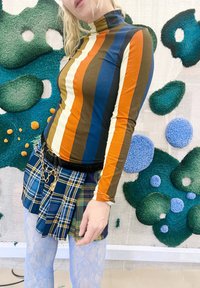 Persona, kas valkā krāsainu svītrainu turtleneck, plānas plīša svārkus ar ķēdes jostu un gaiši zilas rakstainas zeķes priekšā pie teksturētas zaļas un zilas sienas mākslas.