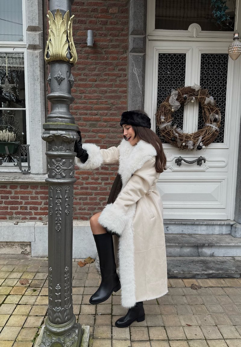 Manteau beige clair avec bordure en fausse fourrure blanche, gants en cuir noir et bottes noires montant aux genoux. Debout à côté d'un lampadaire décoratif.