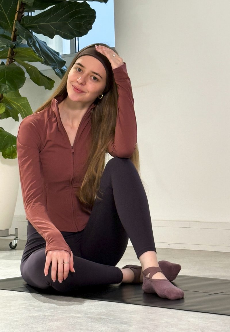 Athletic outfit featuring a zip-up, long-sleeve maroon top and dark leggings, seated on a black mat, with purple socks on feet.