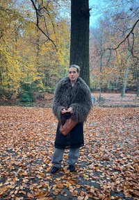 Femme en manteau marron duveteux tenant un sac à main marron, debout sur des feuilles d'automne près d'un grand arbre dans une forêt aux feuillages d'automne colorés.