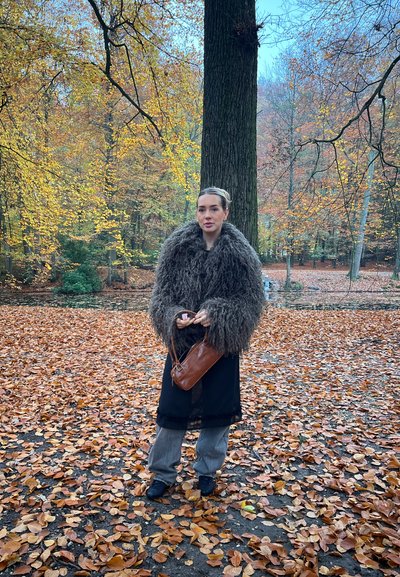 Mujer con abrigo marrón esponjoso sosteniendo un bolso marrón, de pie sobre hojas de otoño junto a un gran árbol en un bosque con follaje colorido de otoño.