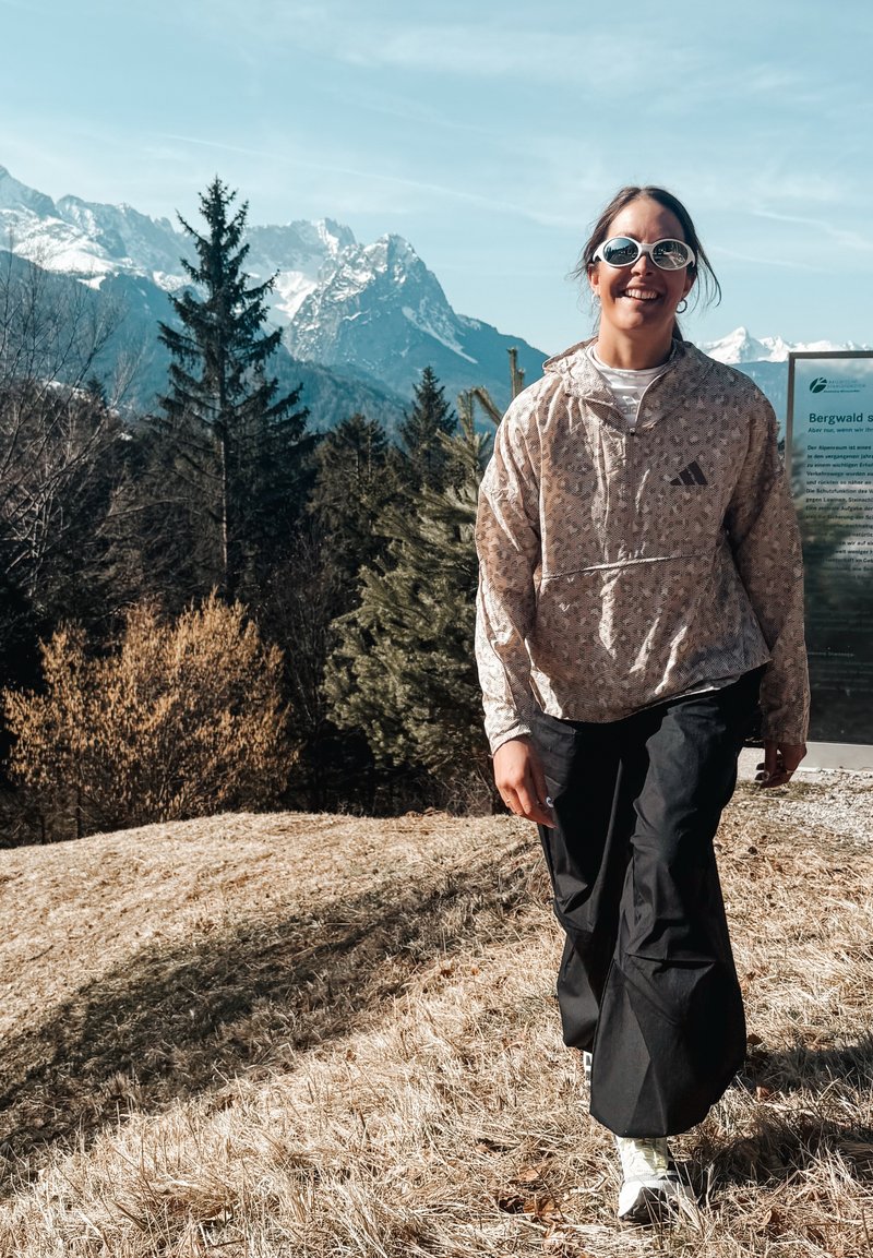 Frau mit Sonnenbrille, die eine leichte Jacke und schwarze Hosen trägt, geht auf trockenem Gras mit schneebedeckten Bergen und Kiefern im Hintergrund spazieren.