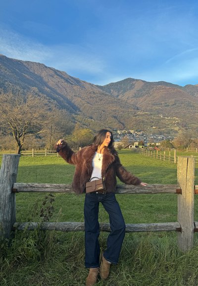 Abrigo de pelo sintético en marrón, blusa blanca y jeans oscuros. De pie junto a una cerca de madera en un campo verde, sosteniendo gafas de sol con un bolso cruzado marrón.