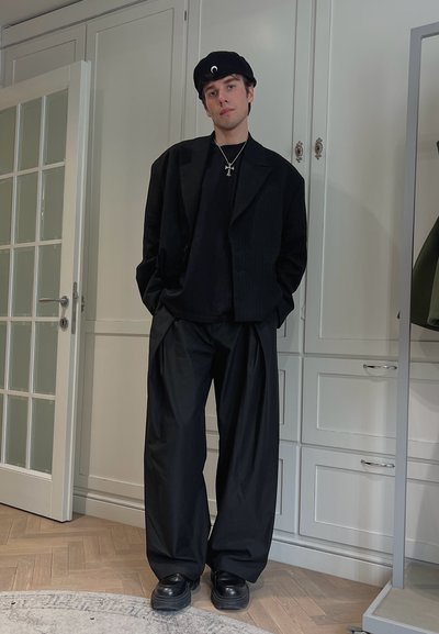 Blazer oversize negro, camiseta negra, pantalones holgados negros, zapatos negros; los accesorios incluyen una boina negra y un collar con cruz plateado.