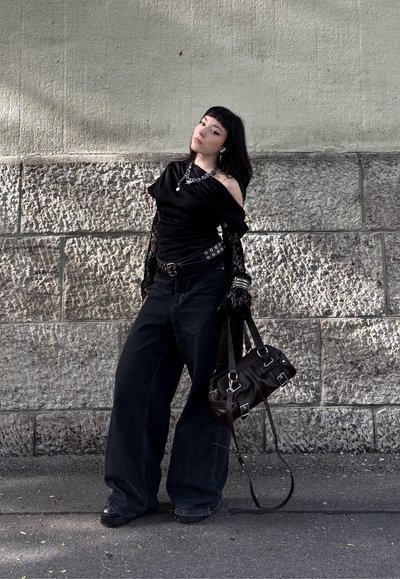 Top negro sin hombros con detalles de encaje, combinado con pantalones vaqueros oscuros sueltos. Bolso negro con herrajes en plata. Fondo de pared de piedra rugosa.