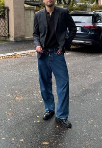 Blazer noir sur une chemise en ribbed noire, associé à un pantalon blue jeans ample et des chaussures noires. Le décor présente un chemin pavé et des véhicules en arrière-plan.