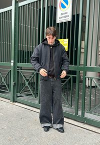 Veste bomber noire portée sur un sweat à capuche noir, associée à un pantalon en denim noir à jambes larges. Comprend une poche zippée et des ourlets effilochés. Portée avec des chaussures noires.