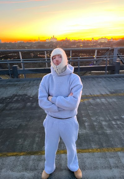 Sudadera y pantalones de chándal de algodón gris claro con capucha y cubrebocas de punto. De pie en una azotea con el horizonte al atardecer de fondo.