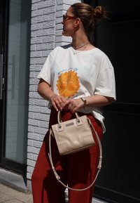 Κρέμα μπλούζα με γραφικό ήλιου, συνδυασμένη με παντελόνι σε ρώσσο χρώμα με φαρδιά ποδόγυρο. Το άτομο κρατά μια μικρή, ανοιχτό καφέ τσάντα χειρός με λαβές και ιμάντα ώμου.