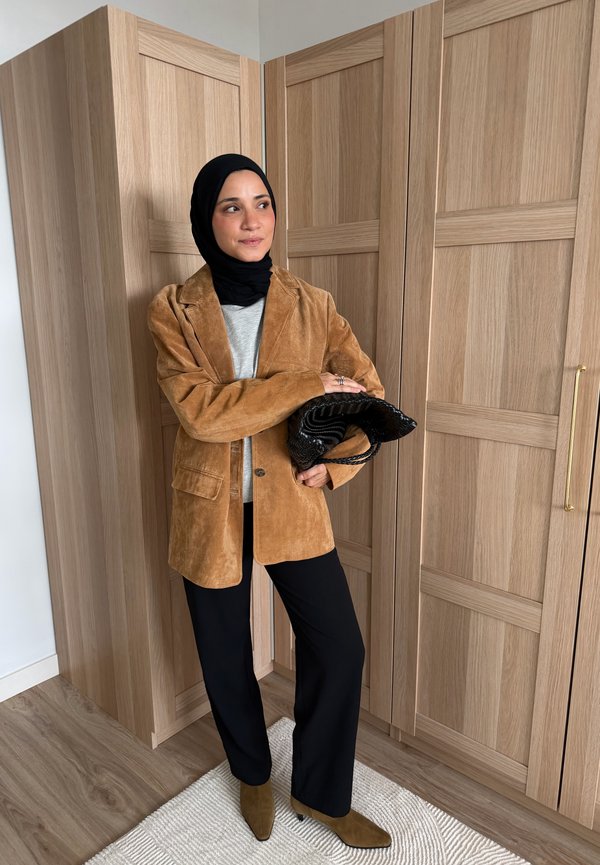 Woman in black hijab, tan suede blazer, black pants, and tan boots holding a black textured clutch, standing near wooden wardrobe.