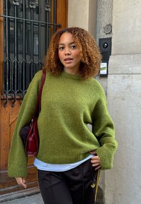 Grønn strikkegenser med avslappet passform, ribbestrikkede mansjetter og rund hals, kombinert med svarte bukser med gule striper. Burgunderfarget veske med dusk.