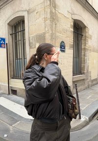 Personne avec une queue de cheval foncée et des lunettes, portant une veste noire et un sac camouflage, debout à un coin de rue en ville.