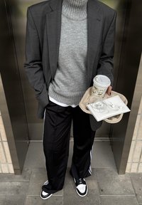 Grau Wollrollkragenpullover kombiniert mit einem dunklen Blazer, schwarzen Jogginghosen mit weißen Streifen und schwarz-weißen Sneakern. Hält eine Kaffeetasse und eine Tasche.