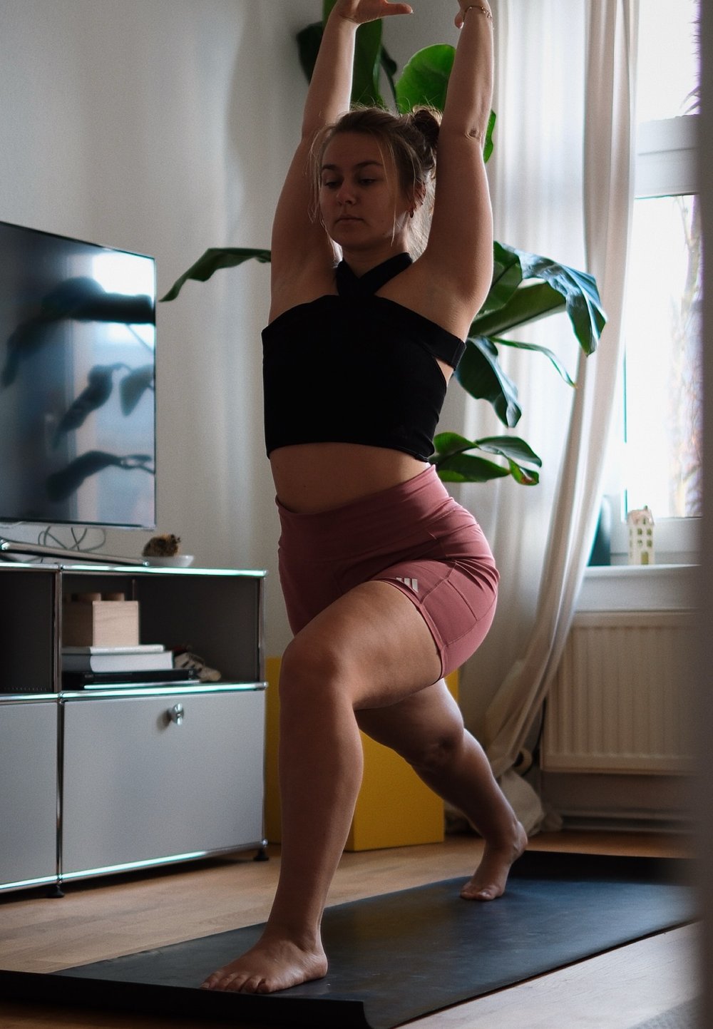 Mulher com um top halter preto e calções cor-de-rosa a executar uma pose de ioga num tapete preto, com plantas em vasos e uma televisão ao fundo.