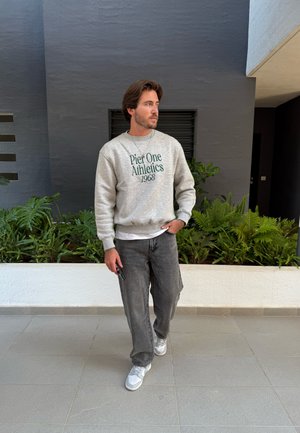 Sudadera gris con la inscripción "Pier One Athletics 1968" en verde. Un par de jeans grises de corte holgado y zapatillas blancas. De pie al aire libre.